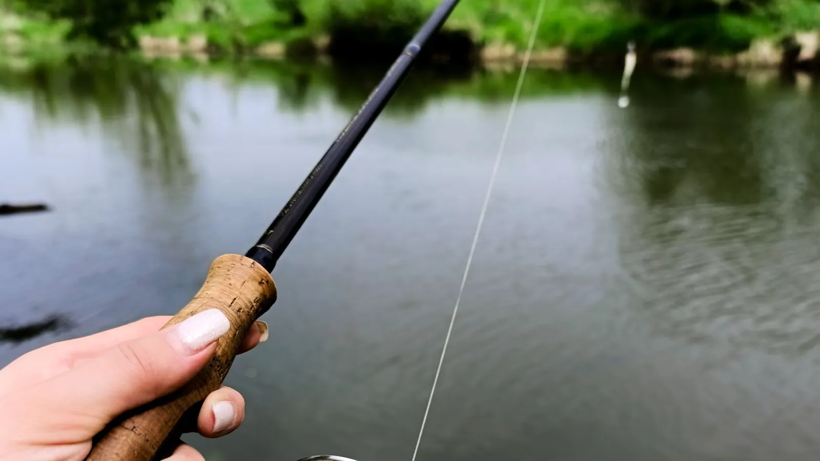 Hand hält eine Spinnrute mit Korkgriff an einem ruhigen Fluss