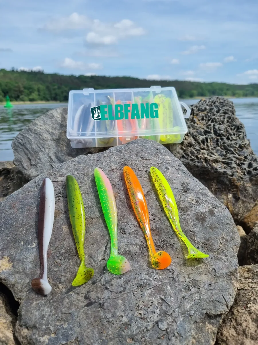 ELBFANG Zander Gummifisch-Set: Köderbox und fünf Gummifische in Weiß-Braun, Grün, Pink-Grün, Orange und Gelb auf einer Steinpackung am Fluss